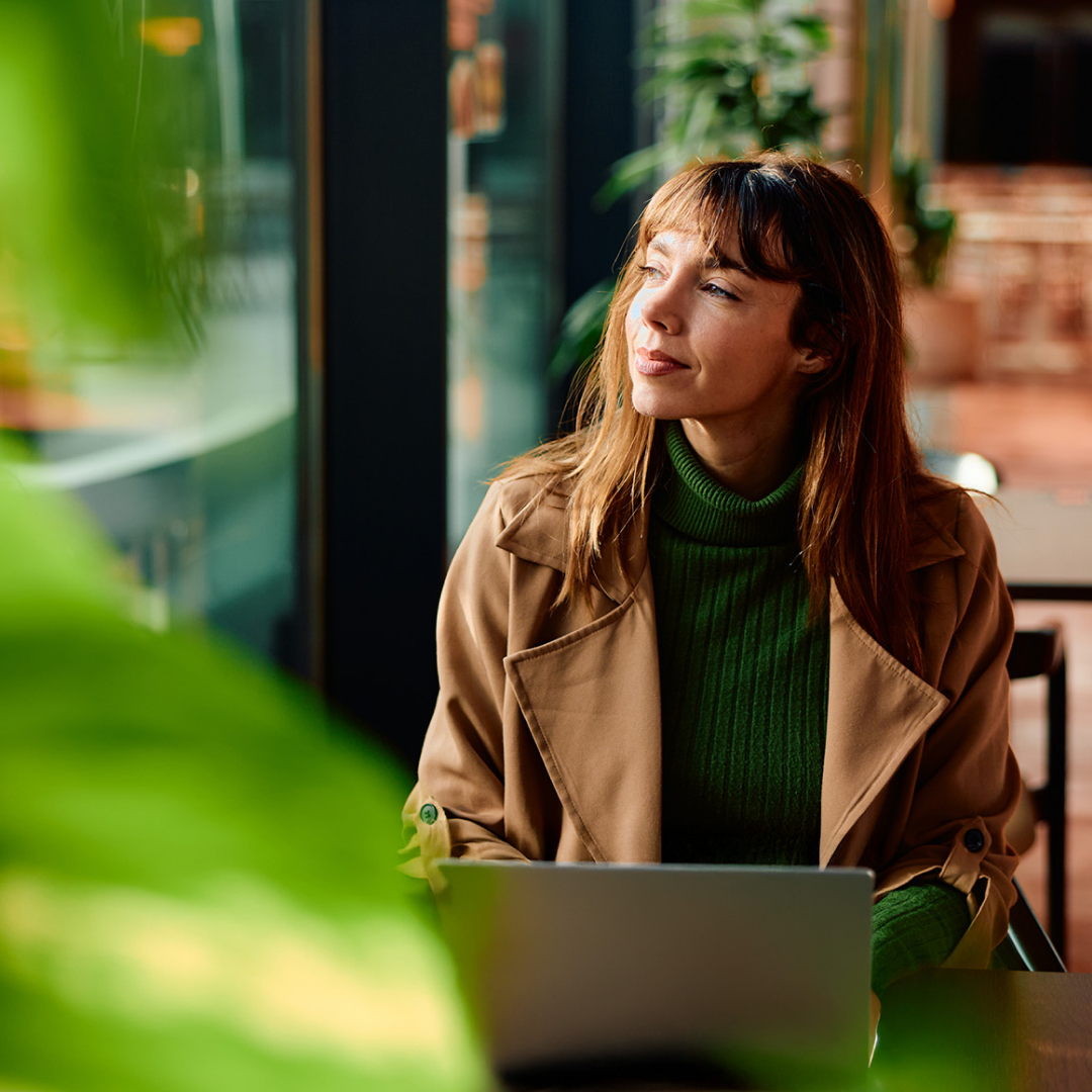 Person working on a laptop in a bright, modern office with large windows and greenery.