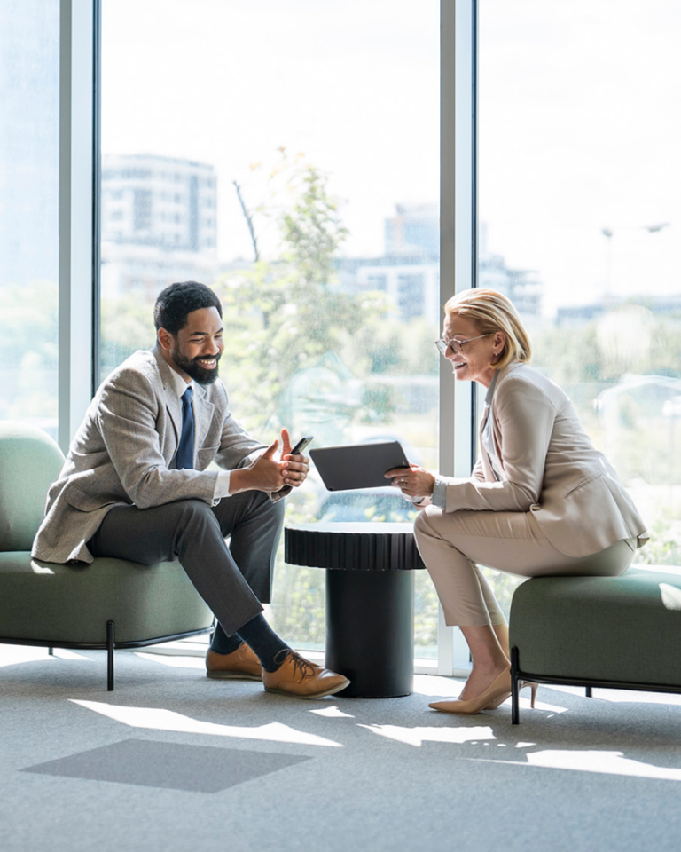 Two professionals meeting in a bright office lounge, reviewing information on a tablet and smartphone.