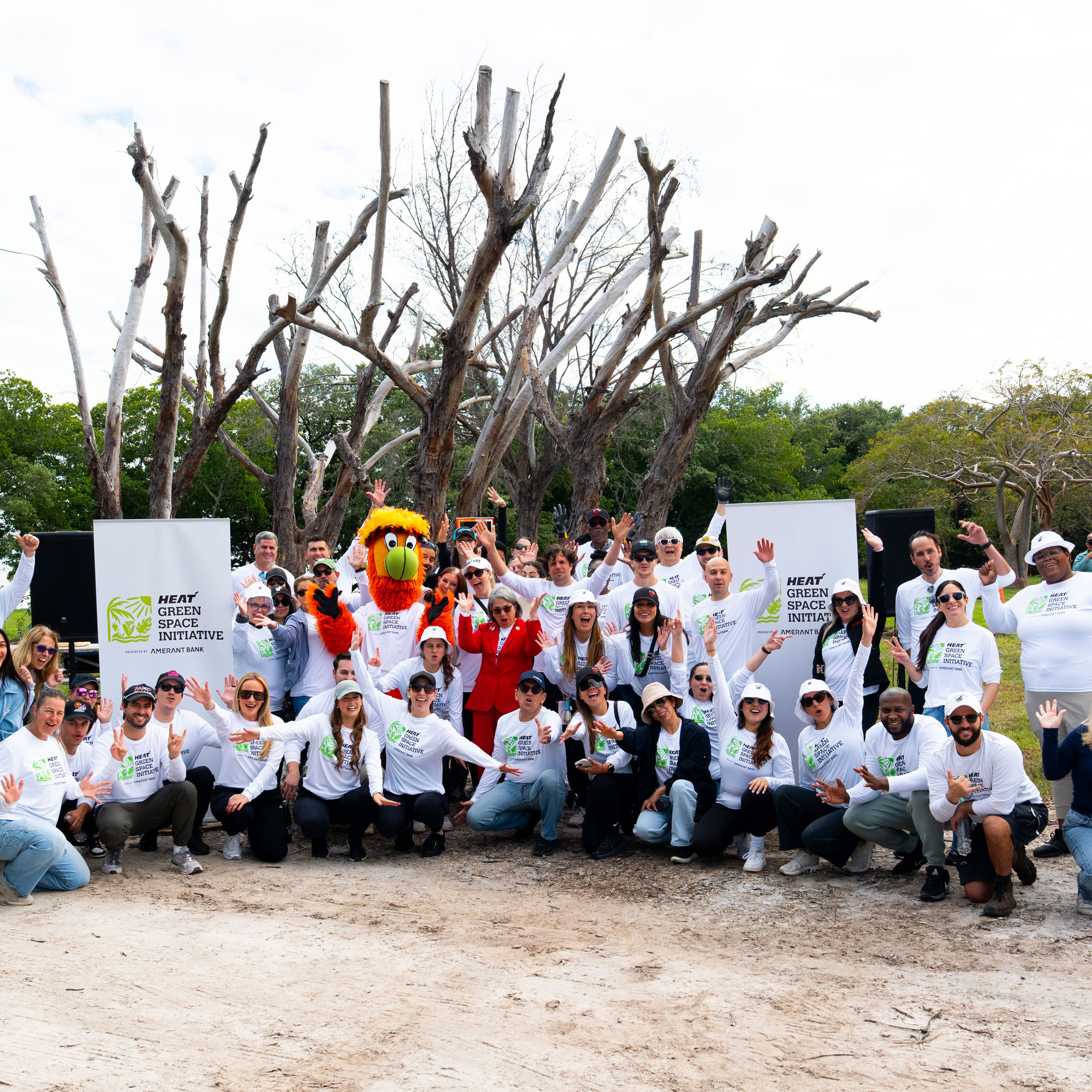 Amerant team members smile with Burnie, the Miami Heat mascot, in front of a tree and in between two banners for the HEAT Green Space initiative event