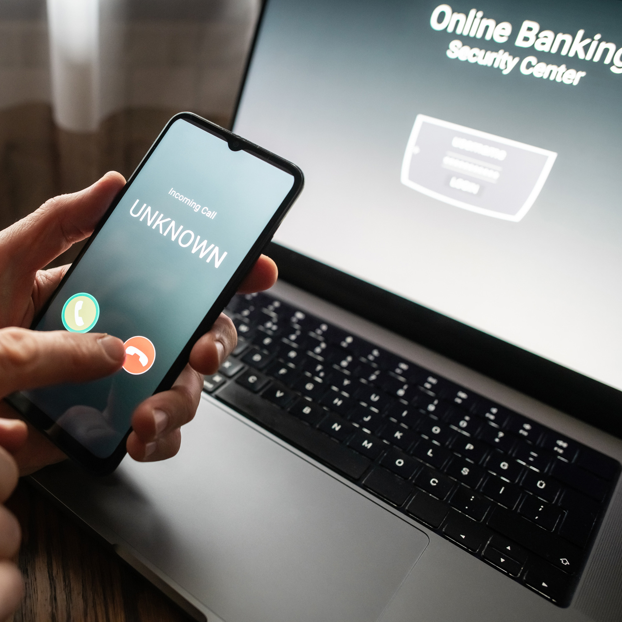 person holding phone up with an unknown number calling and the online banking app on the computer screen behind the phone.