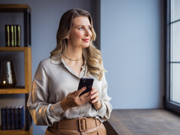 A woman stands indoors near a large window, holding a smartphone with both hands. She wears a light-colored blouse and a tan belt, with a delicate necklace and earrings. Soft natural light from the window illuminates her face as she looks out thoughtfully. A wooden shelf with decorative items and books is visible in the background.