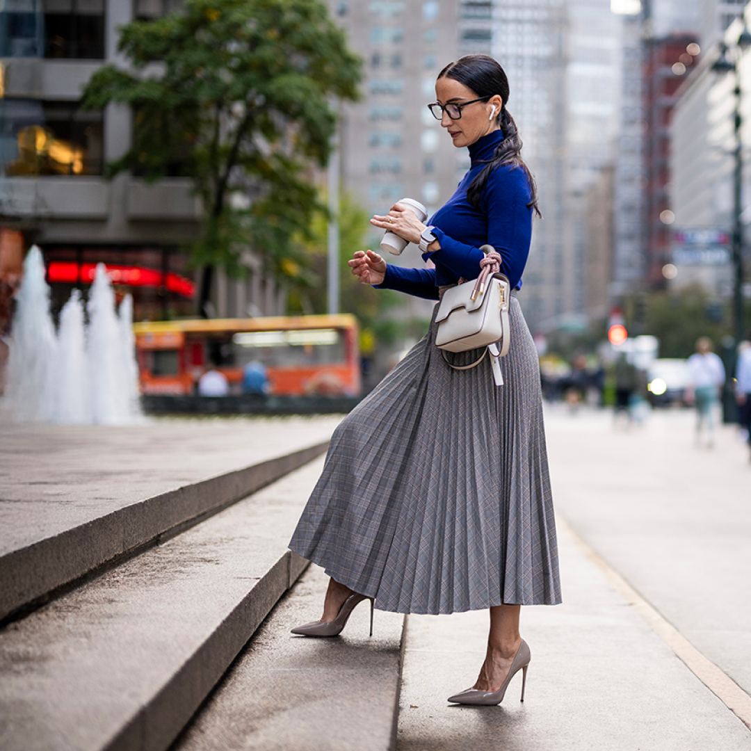 A person walks along a city sidewalk carrying a coffee cup and handbag, with office buildings and a fountain in the background.