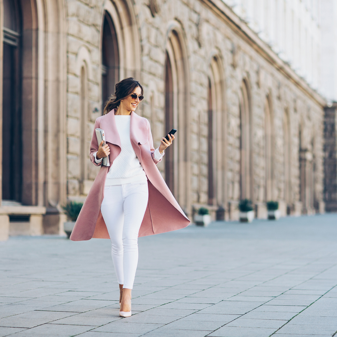 A person walks along a city street beside a historic stone building while looking at a mobile phone.
