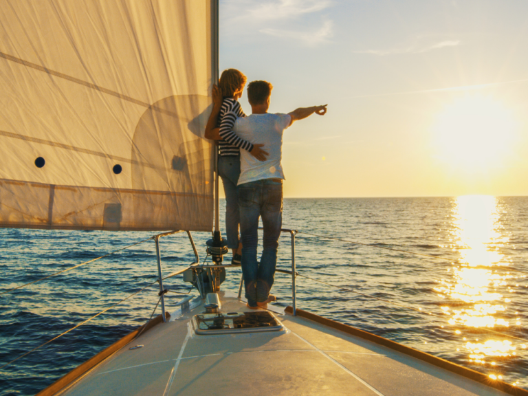 Two people stand on a sailboat at sunset, looking out over the open water and horizon.