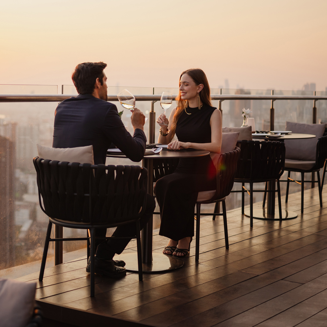 Two people seated at an outdoor rooftop restaurant raise glasses while dining with a city skyline in the background.
