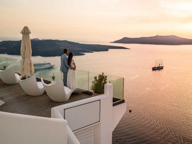 Two people stand on a terrace overlooking a calm sea and island landscape at sunset, with boats visible in the water.