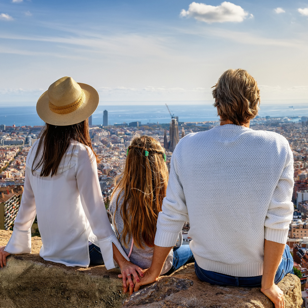 Three people sit on a stone ledge overlooking a dense cityscape with the ocean in the distance. One person wears a straw hat and light blouse, another sits in the center with long hair, and a third wears a light sweater. The person on the left and the person in the center hold hands. The scene is viewed from behind, with buildings, a crane, and a cloudy sky visible across the horizon.