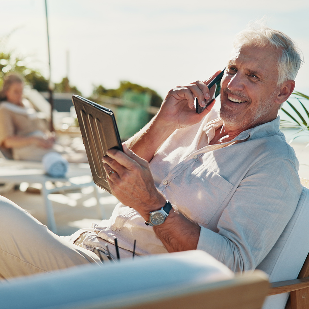 A person relaxes outdoors while talking on a mobile phone and holding a tablet, with lounge seating in the background.