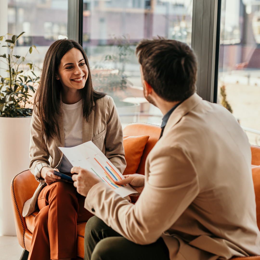 Two people seated in a modern office setting review documents and discuss information during a meeting.