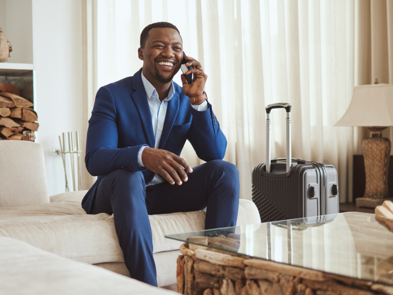 Person in a blue business suit sitting in a hotel lounge, talking on a mobile phone with a rolling suitcase nearby.