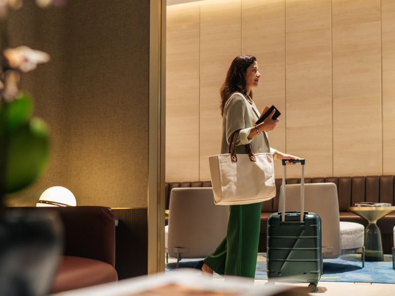 Traveler pulling a rolling suitcase through a hotel lounge while carrying a tote bag and holding a passport.