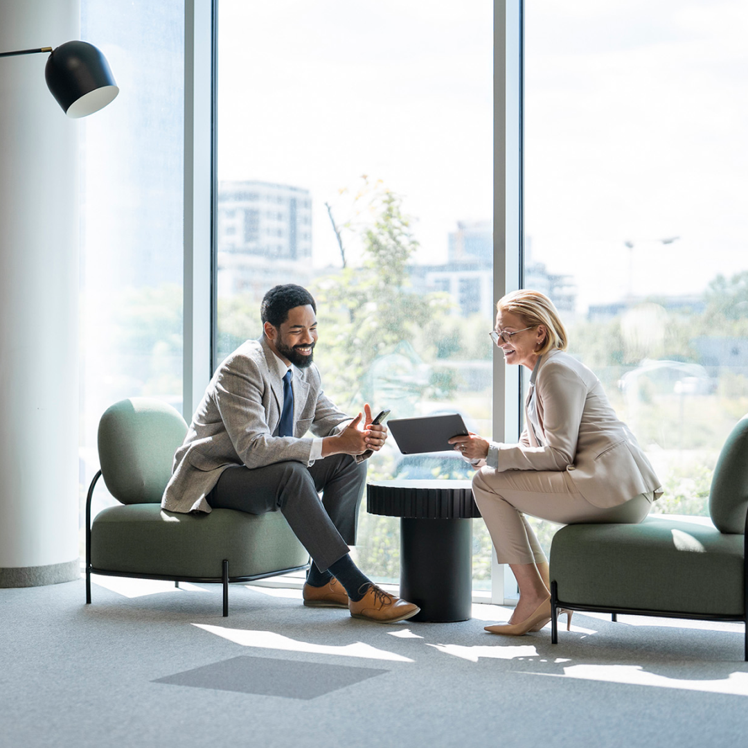 Two professionals meeting in a bright office lounge, reviewing information on a tablet and smartphone.