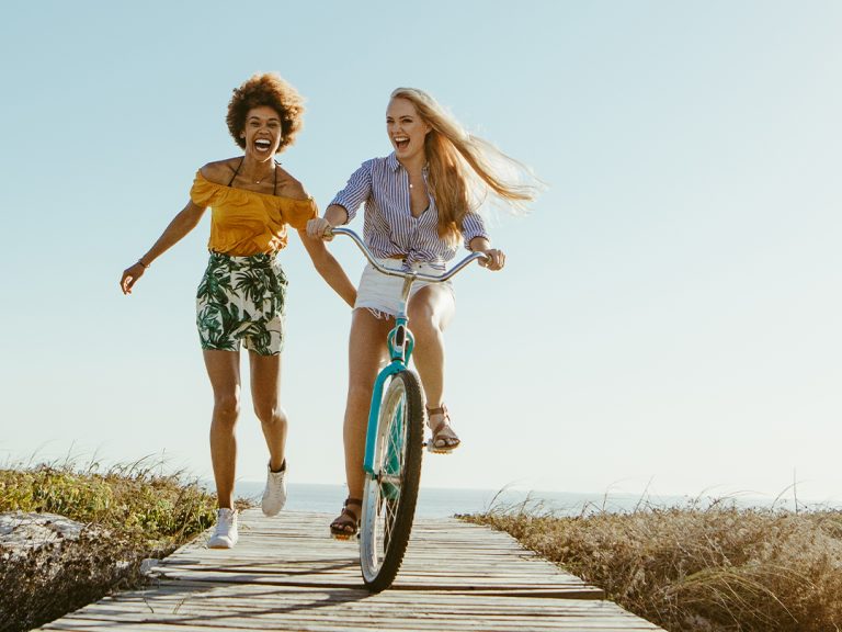 Two people enjoying a sunny coastal boardwalk, with one riding a bicycle and the other walking beside them.