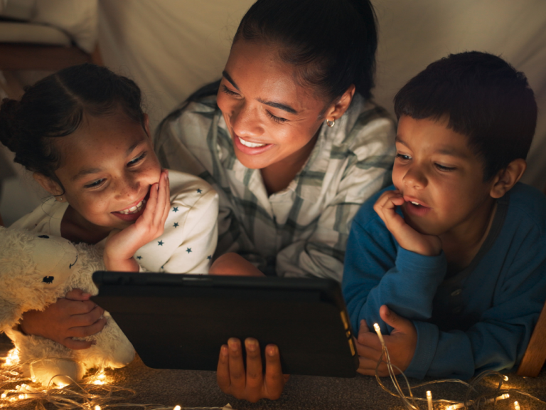 Adult and two children gathered around a tablet inside a blanket fort with warm string lights.